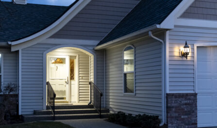 A suburban home’s front porch with a real estate lockbox attached to the railing of the steps is illuminated at evening.