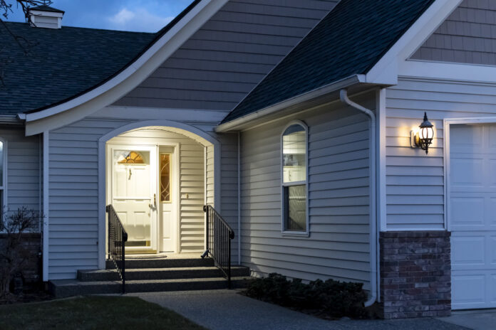 A suburban home’s front porch with a real estate lockbox attached to the railing of the steps is illuminated at evening.